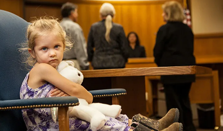 A young girl sits in a courtroom holding a stuffed toy while adults talk in the background near the judge's bench.