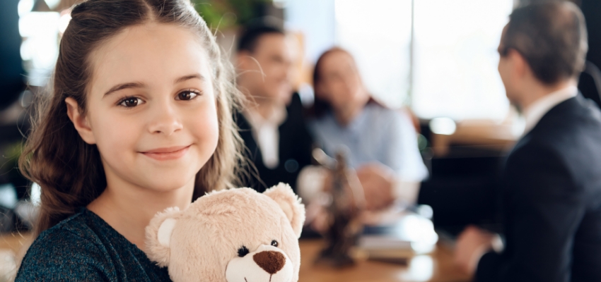 Smiling girl holding a teddy bear in a solicitor’s office, with a lawyer and two adults seated at a table behind her, representing the role of an independent children’s lawyer in parenting matters