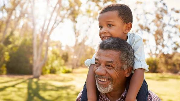 Older man smiling with a young boy on his shoulders in a sunlit park, representing kinship care and child protection lawyer support
